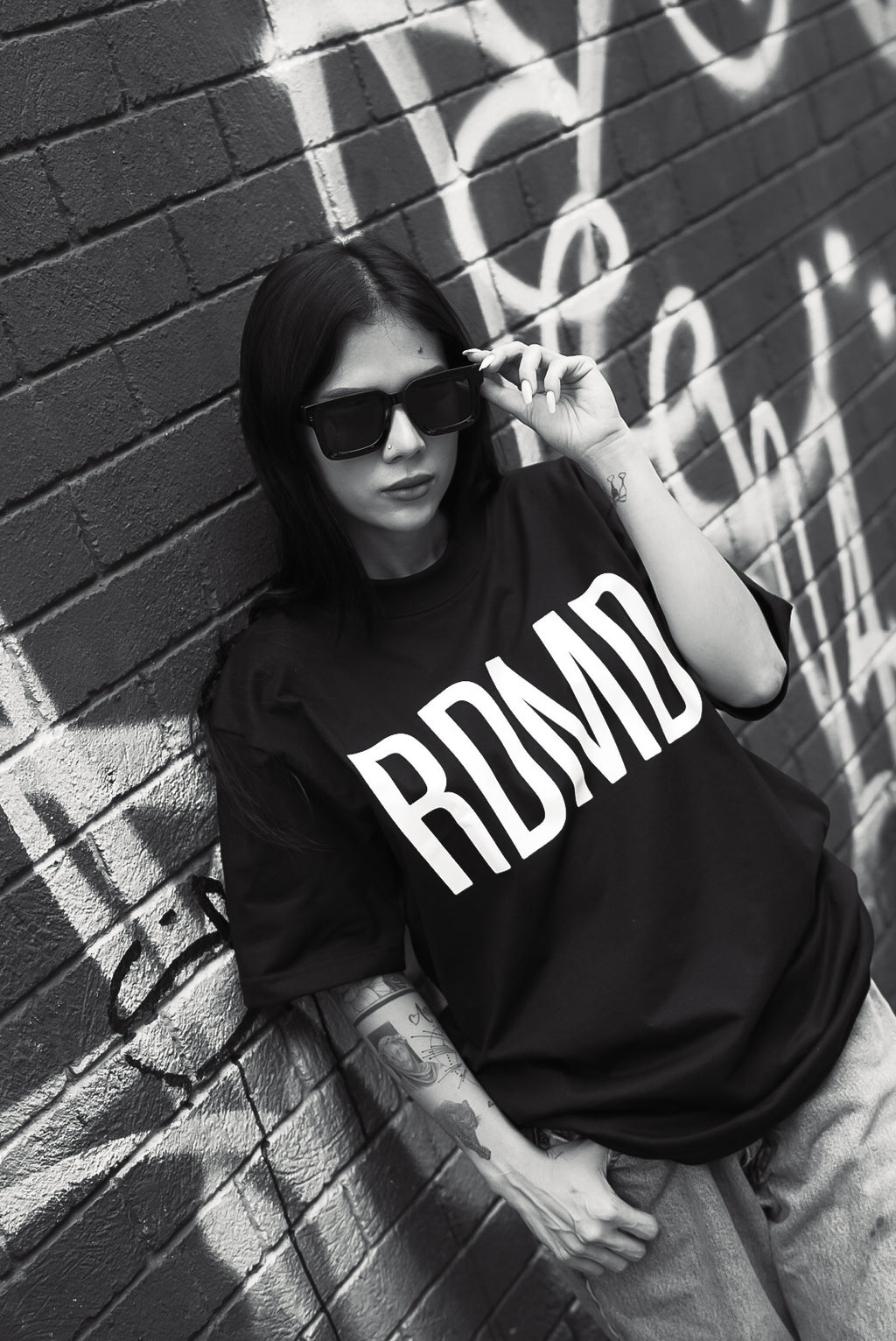Person wearing a black t-shirt with 'RDMD' text, leaning against a graffiti-covered wall.