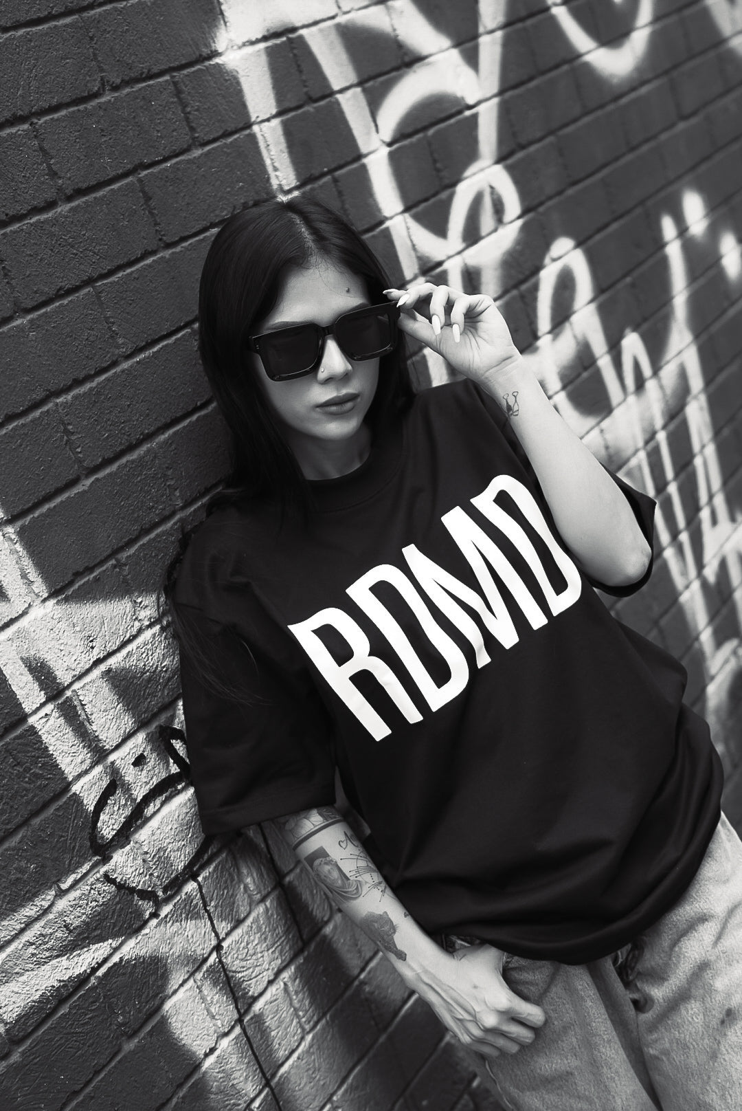 Person wearing a black t-shirt with 'RDMD' text, leaning against a graffiti-covered wall.